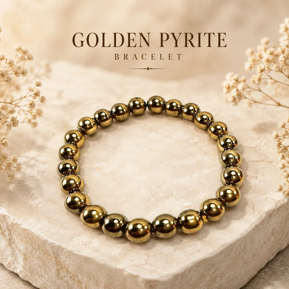 A golden pyrite beaded bracelet arranged in a circle on a light beige stone slab, with dried floral accents and the text 'GOLDEN PYRITE BRACELET' above it.