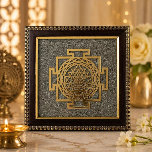 A framed decorative art piece featuring a gold Sri Yantra symbol on a textured gray background, enclosed in a dark brown ornate frame. The frame is displayed on a marble surface with warm bokeh lights and decorative flowers in the background.